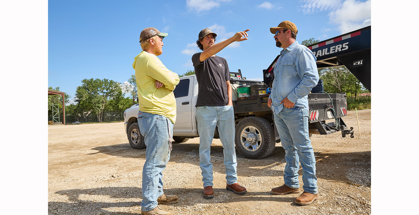 Three men stand talking in a gravel lot near a white pickup truck and a trailer; the man in the center gestures and points into the distance.